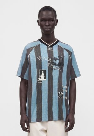 Mand iført en løs blå og sort stribet skjorte med "Venice Beach" tekst og grafiktryk, kombineret med hvide bukser, stående mod en ensfarvet baggrund.