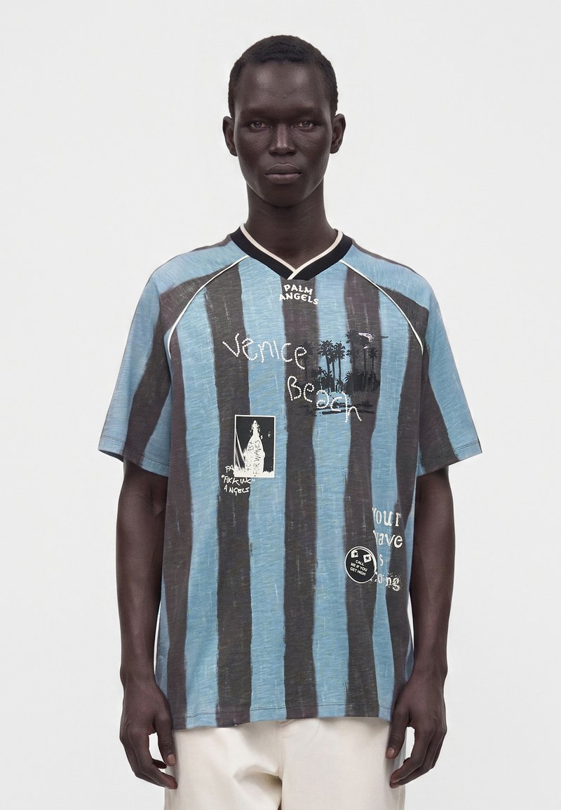 Man wearing a loose blue and black striped shirt with "Venice Beach" text and graphic prints, paired with white pants, standing against a plain background.
