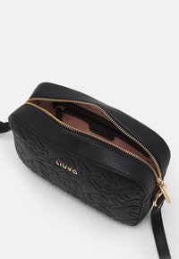Sort tekstureret syntetisk håndtaske med guld lynlås, præget logo og en enkelt justerbar strop. Indersiden har en ferskenfarvet foring.