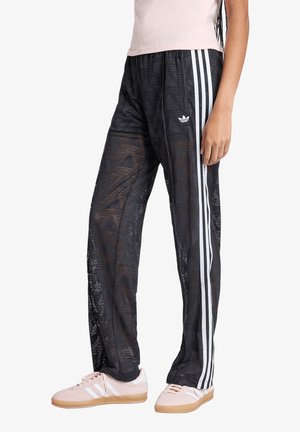 Persoon draagt zwarte Adidas trainingsbroek met witte strepen en logo's, gecombineerd met lichtroze Adidas sneakers met witte strepen.