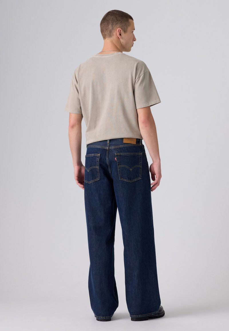 Homme portant une chemise beige à manches courtes et un jean large bleu foncé, debout de dos sur un fond gris uni.