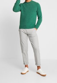Grüner gestrickter Pullover, hellgraue Hose und weiße Sneaker mit beige-samtenen Sohlen. Der Pullover hat einen runden Halsausschnitt und eng anliegende Bündchen.