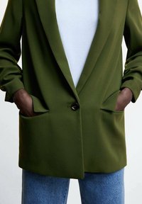 Veste olive avec revers crantés, fermeture par un seul bouton et poches latérales visibles. Portée sur un haut blanc côtelé et un jean bleu.