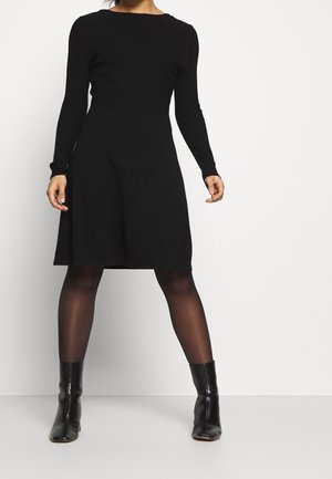 Frau trägt ein schwarzes langärmeliges Strickkleid, transparente schwarze Strumpfhosen und schwarze Knöchelstiefel, steht vor einem schlichten weißen Hintergrund.
