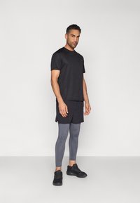 Vyras stovintis dėvintis juod�ą sportinį marškinėlį, juodus šortus, pilkus legingus ir juodus sportbačius ant šviesiai pilko fono.