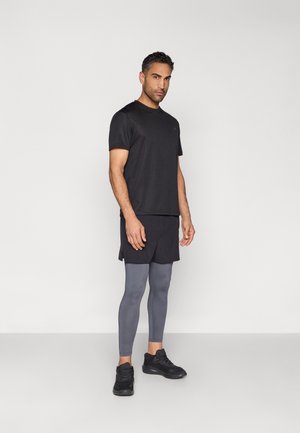 Férfi áll, fekete sportpólót, fekete rövidnadrágot, szürke leggingset és fekete edzőcipőt visel, világosszürke háttér előtt.