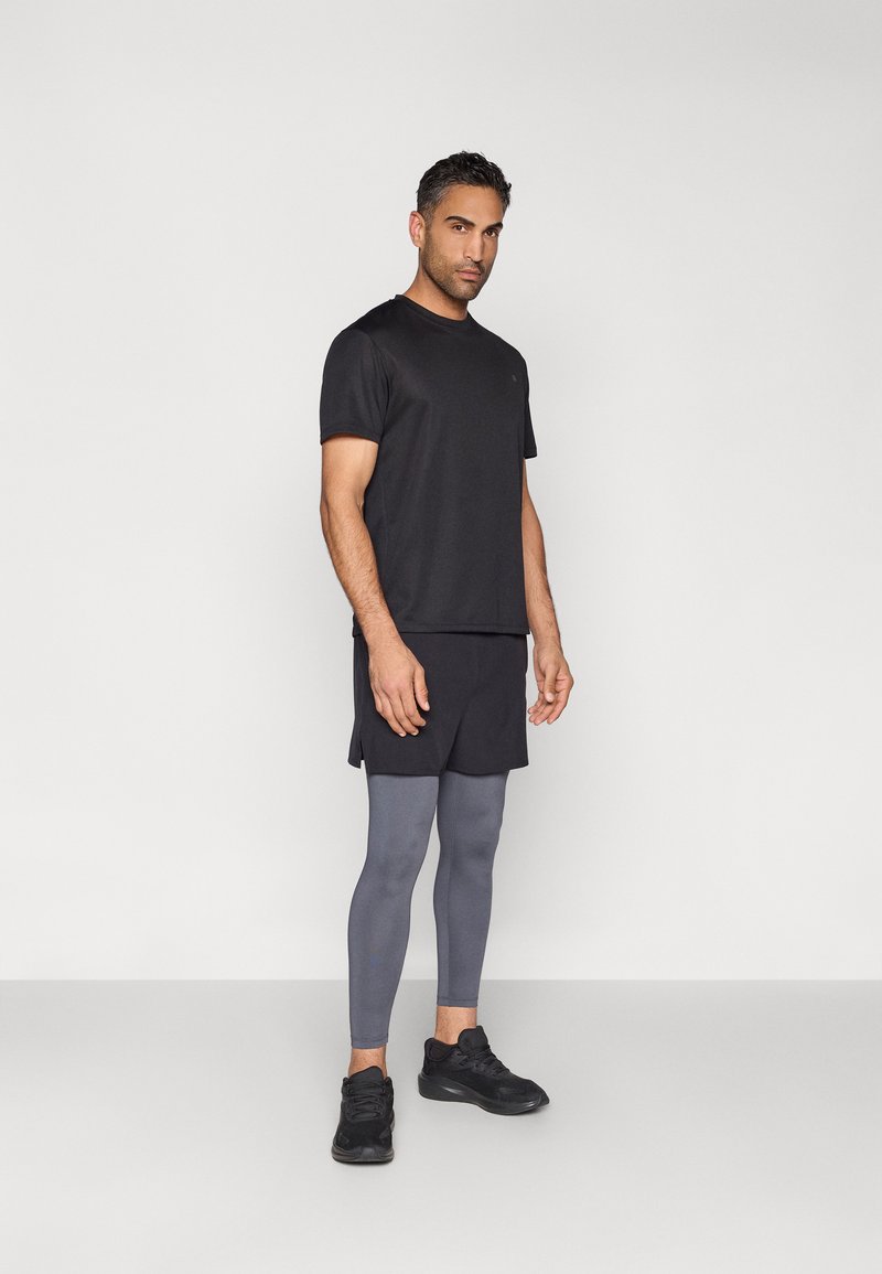 Vyras stovintis dėvintis juodą sportinį marškinėlį, juodus šortus, pilkus legingus ir juodus sportbačius ant šviesiai pilko fono.