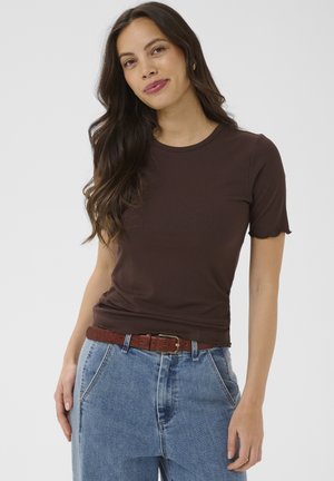 Giovane donna con lunghi capelli scuri, indossa una maglia marrone a maniche corte a coste, jeans blu e una cintura marrone, in piedi davanti a uno sfondo semplice.