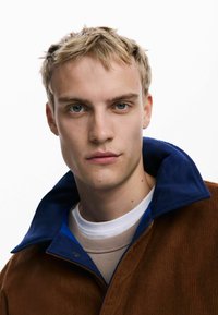 Braune Cordjacke mit blauem Kragen und blauen Akzenten, über einem hellbeigen Hemd getragen. Glatte Textur, minimalistisches Design.