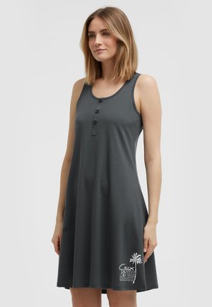 Frau trägt ärmelloses dunkelgraues Kleid mit Knöpfen vorne und weißer Palme sowie Text "THE BEACH" nahe dem Saum auf der linken Seite.