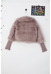 Veste courte en fausse fourrure mauve clair avec poignets allongés côtelés suspendue à un mur blanc, avec un tableau de calibration des couleurs au-dessus.