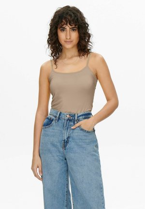 Donna con capelli ricci che indossa una canottiera beige e jeans blu a vita alta, in piedi con una mano in tasca su uno sfondo bianco.