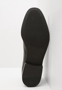 Černá kožená podešev obuvi s texturovaným gripem, mírně zaobleným tvarem, raženým logem a vyztuženou oblastí paty.