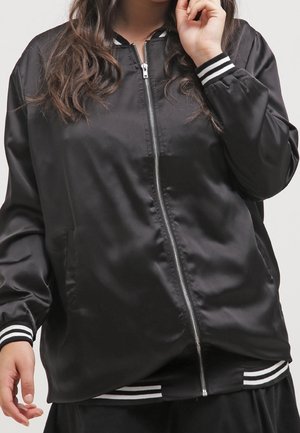 Femme portant une veste bomber satinée noire avec des poignets et une ceinture côtelés à rayures blanches, partiellement zippée, main près du visage et du côté.