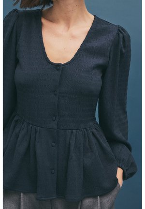 Blouse bleu marine texturée avec col en V, manches longues bouffantes et silhouette péplum. Dotée d'une fermeture à boutons sur le devant et d'un léger froncé.