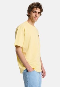 Jeune homme aux cheveux bouclés portant un T-shirt ample jaune clair avec un petit motif sur la poitrine et un jean bleu clair, main dans la poche.