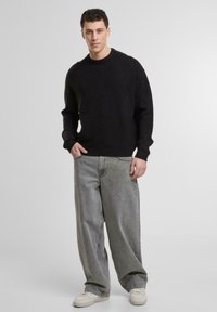 Pull noir texturé à col rond, associé à un jean gris à coupe décontractée et des baskets blanches, se tenant contre un fond clair.