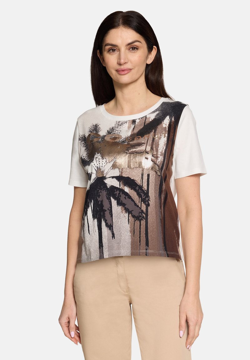 Femme aux cheveux longs et foncés portant un T-shirt blanc à manches courtes avec un motif floral abstrait marron et noir, et un pantalon beige.