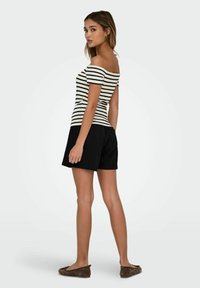 Gestreiftes Off-Shoulder-Oberteil in Schwarz und Weiß, kombiniert mit schwarzen Shorts. Der Stoff wirkt glatt und hat eine taillierte Silhouette. Legere Ballettschuhe.