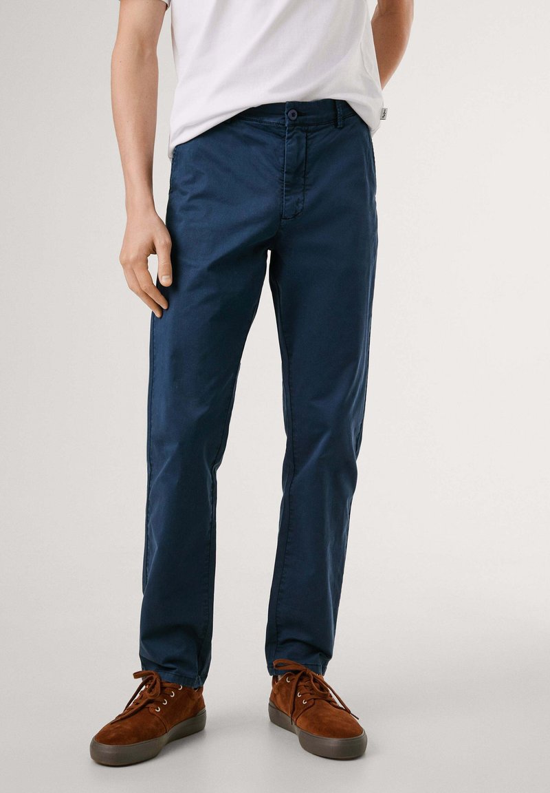 Pantalones de algodón azul marino con textura suave, diseño de pierna recta, cierre de botón delantero y ligero brillo. Combinados con zapatillas deportivas de ante marrón.