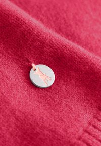 Ronde metalen bedel met een roze honden silhoutte, bevestigd aan een zachte, gestructureerde rode stof. De stof heeft een vlezige uitstraling en een subtiele glans.