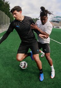 Zwei Fußballspieler in Aktion auf einem grünen Feld. Der eine trägt ein schwarzes, gemustertes Shirt und der andere ein weißes Shirt, beide mit Puma-Logos.