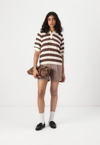 Polo shirt en tricot rayé crème et marron, associé à un short marron. Des chaussures noires et des chaussettes blanches complètent le look. Sac à main marron à la main.