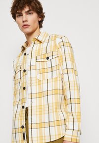 Chemise à carreaux jaunes et blancs en tissu léger, dotée d'une fermeture à boutons, de deux poches poitrine et d'accentuations en noir sur les boutons.