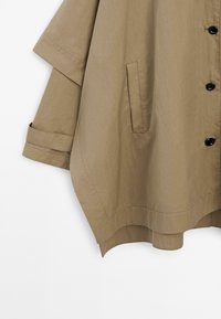 Beige leichte Jacke mit lockerer Passform, kurzen Ärmeln, schwarzen Knöpfen und Seitentaschen. Der Stoff hat eine glatte Textur.