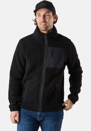 Zwarte fleece jacket met een ritssluiting aan de voorkant, hoge kraag en borstzak. Heeft een gestructureerde stof en zijzakken. Draagt met blauwe spijkerbroeken.