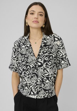 Jeune femme portant une chemise boutonnée à manches courtes noir et blanc avec imprimé léopard et un pantalon noir, accessoirisée de bijoux dorés.