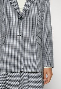Blazer a cuadros con un patrón azul y blanco, cierre de un solo botón, dos bolsillos frontales y solapas con muesca. Detalle de tela texturizada visible.
