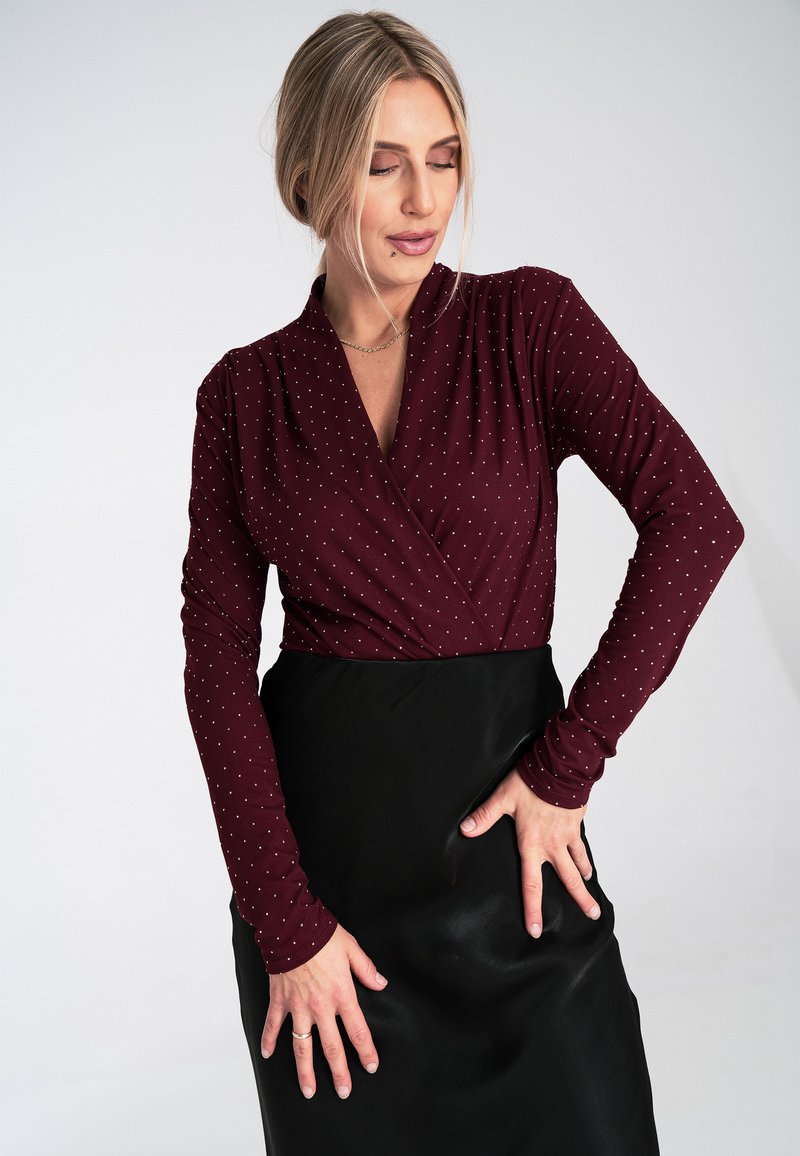 Burgundowa bluzka z długim rękawem w białe kropki, z dekoltem typu kopertowego. Połączona z czarną spódnicą o gładkiej teksturze i dopasowanym kroju.