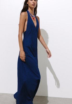 Femme en robe maxi bleue marine sans manches avec un décolleté en V profond et un collier de perles bleu avec un pendentif homard rouge marchant devant un mur blanc.