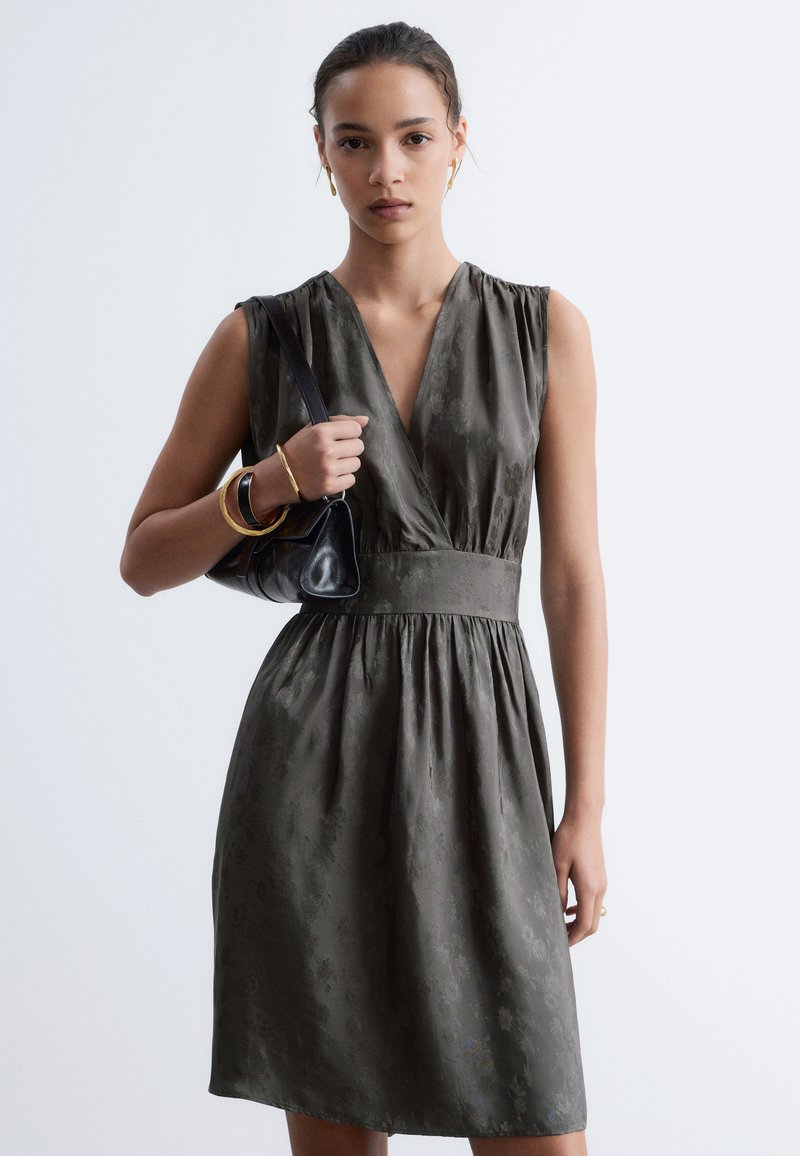 Femme portant une robe sans manches gris foncé avec un motif subtil, tenant un petit sac à main noir, et portant des bracelets et des boucles d'oreilles en or.