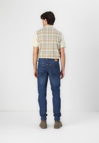 Camisa beige de manga corta con la parte trasera de cuadros, combinada con jeans azules y botas marrones. El conjunto presenta líneas limpias y un ajuste relajado.