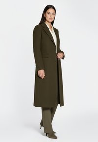 Olivengroene lange jas met een getailleerd silhouet, reverskraag, twee voorzakken en een gestructureerde stof, gecombineerd met een crèmekleurige blouse en bijpassende broek.