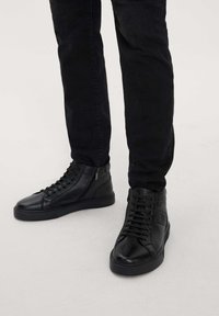 Adidași high-top din piele neagră, cu o textură netedă, ochiuri contrastante și un logo texturat pe partea exterioară. Șireturile se întind pe față.