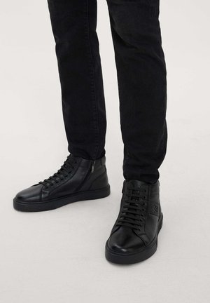 Zapatillas altas de piel negra con una textura suave, ojales en contraste y un logo texturizado en el costado exterior. Los cordones suben por el frente.