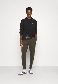 Schwarzer Hoodie mit Kordelzug, olivgrüne Jogginghose und weiße Sneakers mit schwarzen Streifen. Das Outfit wird durch eine schwarze Bauchtasche an der Taille ergänzt.