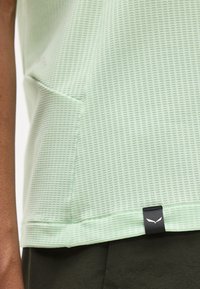 Φωτεινό πράσινο πουκάμισο από υφασμάτινη υφή με μια μικρή μαύρη ετικέτα κοντά στο τελείωμα, φορεμένο πάνω από σκούρο παντελόνι στον βραχίονα και την περιοχή της μέσης ενός ατόμου.