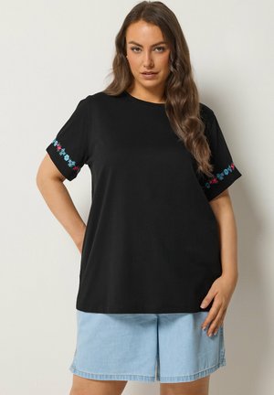 Kobieta ubrana w czarną koszulkę z krótkim rękawem z niebiesko-różowym kwiatowym haftem na rękawach oraz jasnoniebieskie szorty dżinsowe, stojąca na tle białej ściany.