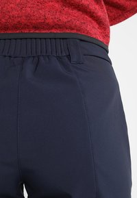 Zbliżenie na osobę w granatowych spodniach z elastycznym pasem i pętlą na pasek, w połączeniu z czerwonym, teksturowanym topem.