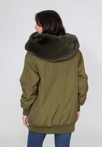 Olijfgroene bomberjack met een pluche grijs bontkraag; beschikt over ribgebreide manchetten en een tailleband, ritssluiting en zijzakken.