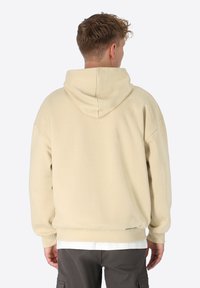 Beige Kapuzenpullover mit lockerer Passform, der über eine weiche Textur und ein Logo-Detail am Saum verfügt. Sichtbare Kängurutasche und Kapuze mit Kordelzug.