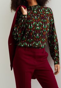 Långärmad blus med hög hals i ett flerfärgat, abstrakt mönster med gröna, bruna och vita nyanser, tillsammans med bourgogne-färgade skräddarsydda byxor.