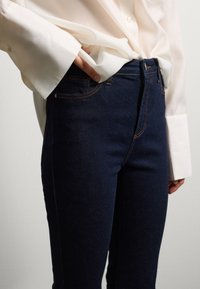 Jeans in denim blu scuro con taglio slim, caratterizzati da dettagli di cucitura arancione, chiusura centrale con bottone e tasche laterali. Abbinati a una camicia bianca.