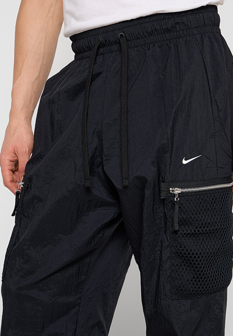 Uomo che indossa pantaloni neri Nike con cintura elastica, stringhe, tasca in rete con zip e logo Nike bianco sulla coscia destra.