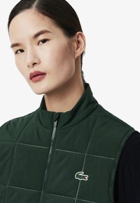Groene gewatteerde vest met hoge kraag, voorzien van subtiele stiksels, een ritssluiting aan de voorkant en een klein Lacoste-logo op de linkerborst.