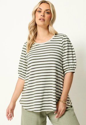 Femme portant une chemise rayée blanche et vert olive à manches bouffantes et un pantalon vert clair, debout devant un fond clair uni.